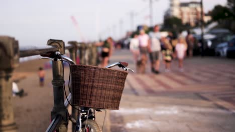 Bicicleta-Antigua-Con-Cesta-Estacionada-En-La-Acera-Durante-La-Puesta-De-Sol,-Gente-Caminando-Detrás