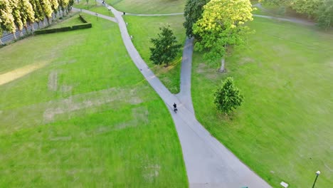 Person-in-black-clothing-cycles-over-the-gray-asphalt-in-Hillsborough-Park-among-the-green-lawns-and-trees-in-the-English-city-of-Sheffield