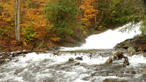 Herbstwaldszene-Mit-Gefallenen-Blättern,-Die-Im-Wind-über-Einem-Kaskadierenden-Wasserfall-Wehen
