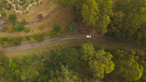 High-altitude-aerial-perspective-tracking-white-vehicle-driving-along-dirt-road-below-tree-canopy