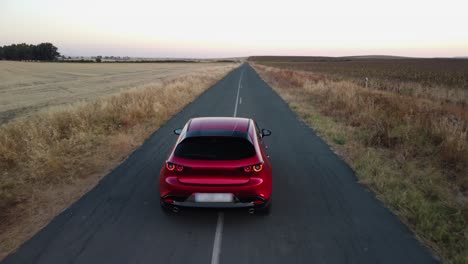 Shiny-new-red-car-drives-slowly-on-narrow-road-through-crop-fields