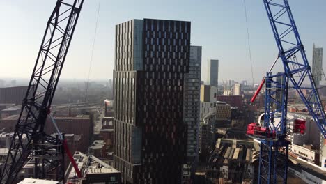 Aerial-drone-flight-between-two-construction-cranes-on-Oxford-Road-in-Manchester-City-and-rising-above-and-over-a-tower-rooftop-to-reveal-a-skyline-of-the-South-Towers-in-the-distance