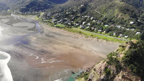 Drone-tilt-up-next-to-Lion-Rock-reveals-charming-Piha-Village,-West-Auckland