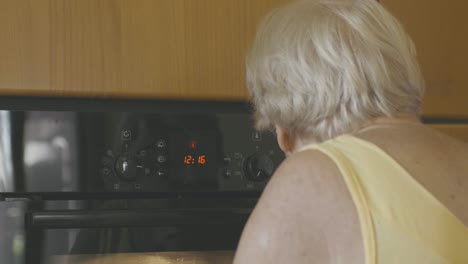 Eine-ältere-Frau-Schaut-Auf-Den-Ofen-Und-Wählt-Aus,-Mit-Welcher-Temperatur-Und-Welcher-Uhrzeit-Ein-Kuchen-Gebacken-Werden-Soll
