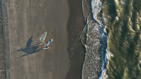Couple-of-windsurfer-leaving-North-Sea-walking-on-sandy-beach-with-kite-at-sunset---aerial-top-down