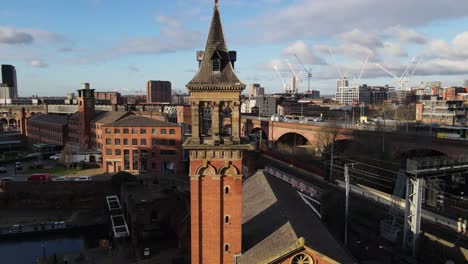 Aerial-drone-flight-over-Deansgate-Congregational-Chapel-in-Manchester-City-Centre-showing-a-tram-passing-by-over-the-viaducts-and-canal-boats-in-Castlefields-Quays