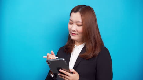 Schöne-Asiatische-Geschäftsfrau-In-Einem-Schwarzen-Anzug,-Der-Ein-Digitales-Tablet-Für-Die-Planung-Von-Online-arbeiten-Mit-Drahtloser-Technologie-Auf-Blauem-Hintergrund-Verwendet