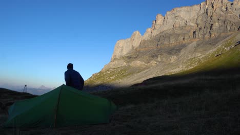 Ein-Mann-Steht-An-Einem-Frischen-Morgen-Vor-Seinem-Zelt-Auf-Und-Blickt-Auf-Eine-Sonnenbeschienene-Felswand-In-Den-Französischen-Alpen
