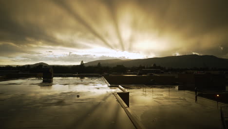 Raindrops-fall-on-the-puddles-formed-on-a-rooftop-as-the-sun-sets-beautifully-behind-distant-mountains,-creating-god-rays-from-the-clouds