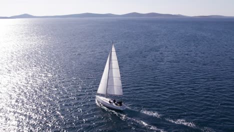 White-sailing-yacht-moving-into-bright-sunshine-reflecting-on-sea-surface,-aerial
