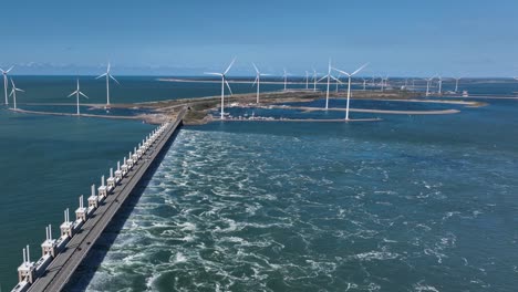 Aerial-flyover-storm-surge-barrier-dam-at-Delta-Works-in-netherlands-with-rotating-offshore-wind-turbines-in-background---Cars-crossing-Zeeland-Bridge