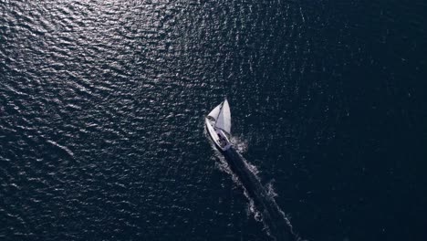 White-sail-boat-cruising-through-blue-sea-with-bright-sunshine-on-surface,-aerial