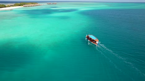 Vista-Aérea-De-La-Película-Del-Barco-Dhoni-De-Maldivas-A-Través-De-La-Bahía-A-La-Isla-Con-Bungalows-Sobre-El-Agua