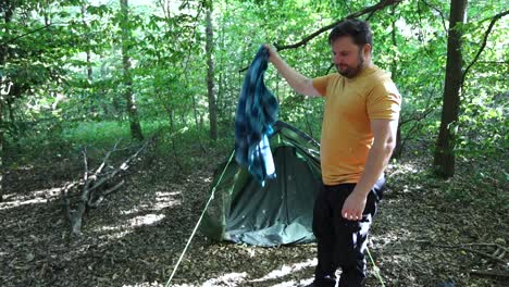 Slow-motion-of-man-tourist-shaking-leaves-from-a-blanket-in-front-of-a-tent-at-forest-campsite