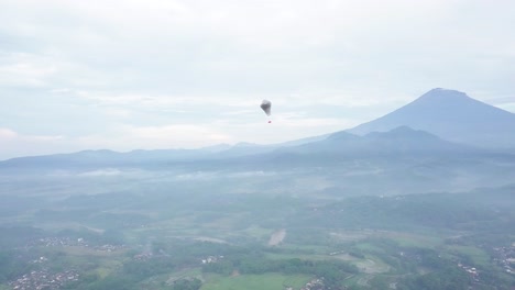 Aerial-tracking-shot-of-INDONESIAN-TRADITIONAL-BALLOON-with-flag-flying-between-clouds-in-rural-mountain-landscape