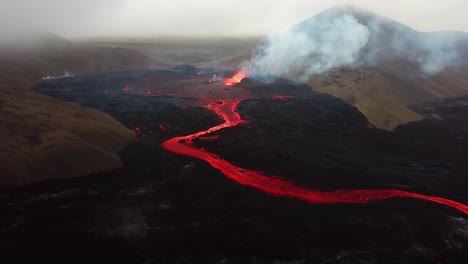 Aerial-landscape-view-of-Fagradalsfjall-volcano-erupting-with-lava-flowing-across-the-Meradalir-valley-floor,-and-smoke-coming-out