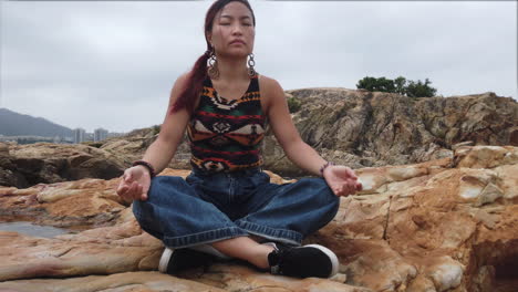 Chica-Meditando-Con-Las-Piernas-Cruzadas-Sobre-Una-Roca-Junto-Al-Mar-Al-Aire-Libre-En-Un-Día-Nublado