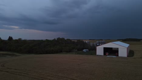 Curved-drone-flight-approaching-a-large-farm-hangar-and-giant-grain-bins-at-nightfall-in-the-remote-countryside-of-north-American-prairies