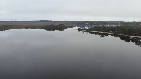 Aerial:-Drone-flying-over-a-crystal-still-lake-towards-a-harbour-full-of-boats-on-a-moody-morning-in-Strahan,-Tasmania