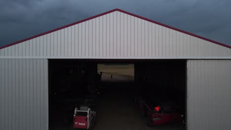 Large,-white-hangar-with-farm-machinery-inside-and-a-person-standing-at-the-other-end-during-nightfall