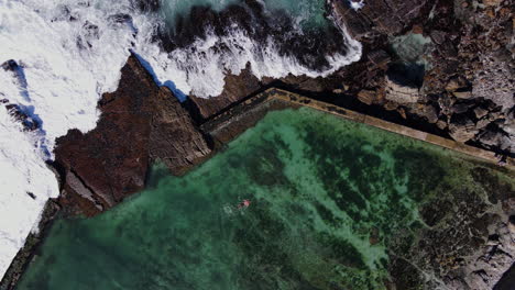 Top-down-aerial-of-man-swimming-in-tide-pool-on-rocky-coast-with-crashing-waves