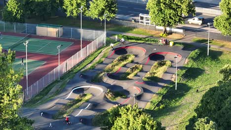 Young-people-on-scooters-and-mountain-bikes-ride-a-hilly-skate-park-next-to-the-tennis-courts-in-Hillsborough-Park-in-Sheffield-on-a-sunny-summer-day