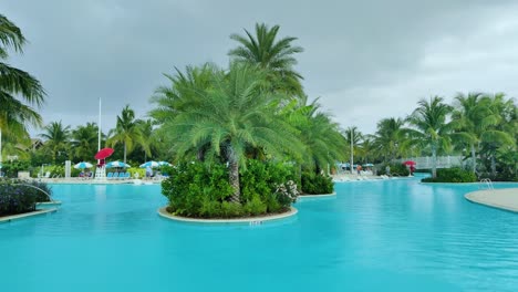 Ein-Riesiges-Schwimmbad-Mit-Palmen-In-Der-Mitte-Eine-Tropische-Insel-Mit-Bewölktem-Und-Regnerischem-Wettervideohintergrund-In-4k-|-Tropeninsel-Mit-Palmen-Bewölkt-Und-Regenwetter-Videohintergrund