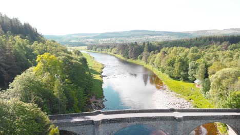 Stone-bridge-across-river-in-highland-nature-of-Scotland,-sunny,-drone
