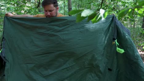 Young-adult-man-packing-up-tent-after-sleeping-in-forest,-handheld-view