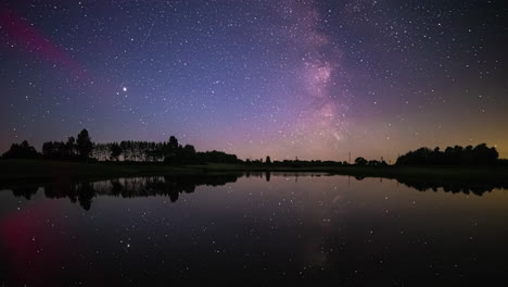 Night-Sky-Full-Of-Stars-Reflecting-On-Calm-Lake-Water-With-Vehicular-Lights-Glowing-In-Distance