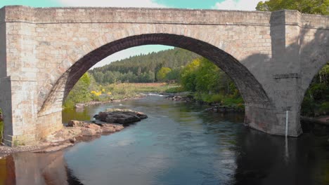 Stone-bridge-arches-over-flowing-highland-river-in-Scotland,-drone