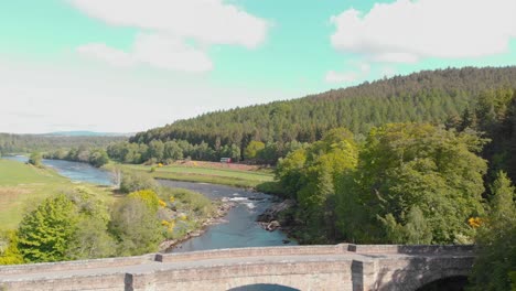 Bus-driving-along-river-with-bridge-in-sunny-scottish-highlands,-drone