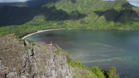 Traveler-couple-stands-on-Crouching-Lion-Rock-in-Hawaii,-drone-view-of-Kahana-bay