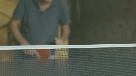 Ping-pong-net-in-the-foreground-and-in-focus,-in-the-background-not-in-focus-a-man-catches-the-ball-with-a-red-paddle-and-plays-with-whoever-is-beyond-the-camera,-slow-motion,-interior-daytime