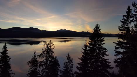 Silhouette-pine-tree-reveals-glassy-reflective-lake-and-mountains,-sunset,-60fps