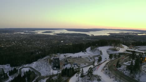 Oslo-city-pull-back-2-with-Norwegian-sea,-Vinterpark-Winterpark-Tryvann-Drone-Push-in-Past-Ski-Jump-at-Sunset-Holmenkollen-ski-jump