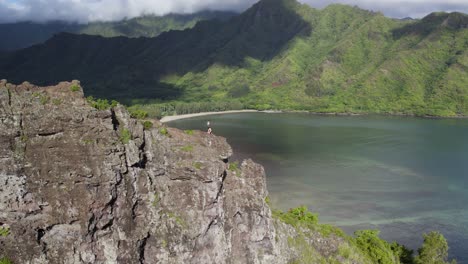 Incredible-aerial-drone-pov-with-natural-parallax-perspective-of-people-on-top-of-dangerous-cliff-on-Crouching-Lion-Hike-with-breathtaking-view-of-Kahana-valley-and-bay,-Hawaii