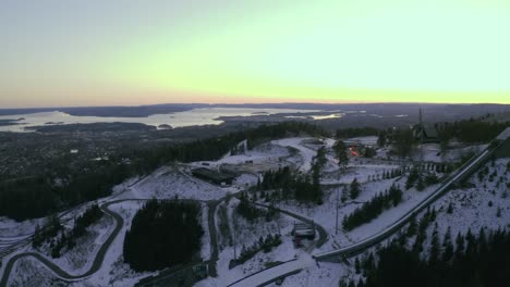 Oslo-city-with-Holmenkollbakken-ski-jump,-Vinterpark-Winter-park-Tryvann-Drone-wrap-around-Past-Ski-Jump-at-Sunset-Holmenkollen