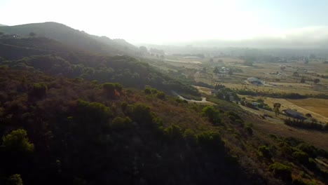 Drone-Disparó-Sobre-La-Colina-Con-Sol-A-Través-De-La-Niebla-Y-La-Granja-En-El-Fondo