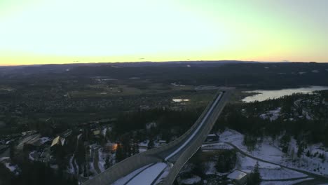 Holmenkollen-ski-jump-drone-pull-back,-Oslo-city-with-Norwegian-sea-in-background,-Vinterpark-Winterpark-Tryvann-with-nordic-cross-country-skiing-at-Sunset