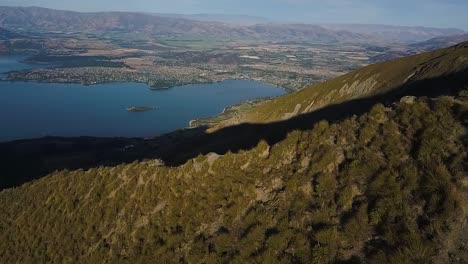 A-drone-shot-moving-down-the-edge-of-a-mountain-reveals-a-vast-valley-and-lake-backdrop