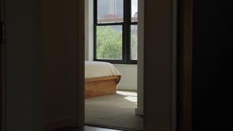 The-corner-of-a-bed-from-outside-the-room-in-the-hallway-with-a-window-in-the-background