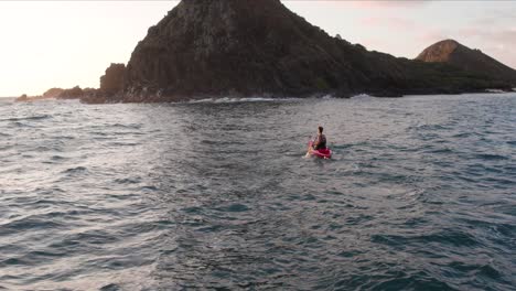Cinematic-drone-back-view-of-solitary-man-paddling-on-red-kayak-toward-Na-Mokulua-Hawaiian-islands