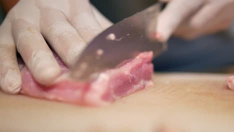 Hand-with-Glove-Use-Knife-to-Cut-Pork-Loin-into-Pieces-on-White-Chopping-Board,-Close-Up