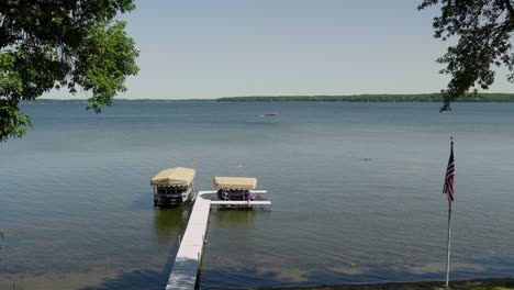 Dock-with-a-pontoon-and-jet-skis-on-a-large-lake-with-an-American-flag-in-the-foreground-and-a-pontoon-driving-in-the-background