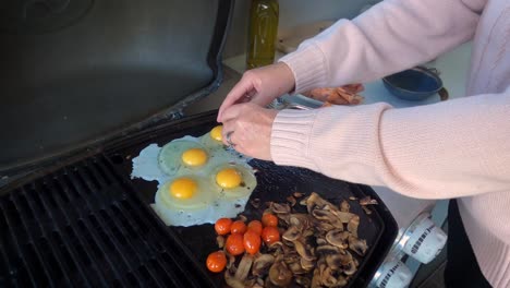 Panning-out-from-a-well-dressed-lady-in-a-pink-sweater,-cracking-eggs-and-frying-them-on-a-modern-home-barbecue