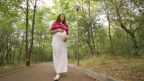 Looking-up-at-pregnant-woman-walking-past-park-lamp-post-on-paved-trail,-slowmo