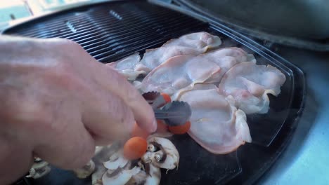 Closeup-of-a-hand,-turning-fried-bacon,-mushrooms-and-tomatoes-with-tongs-on-a-modern-inbuilt-barbeque