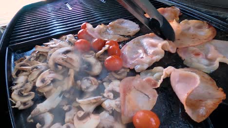 Turning-bacon,-mushrooms-and-tomatoes-with-tongs-on-a-hot-smokey-barbecue