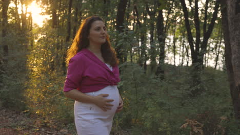 Pregnant-mom-looking-ahead-while-walking-in-forest-as-exercise,-sun-sets-behind
